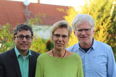 Das Bild zeigt drei Menschen: Links ein Mann mit Brille, in der Mitte eine Frau mit Brille, rechts ein Mann mit Brille. Im Hintergrund sind ein Haus und Bäume zu sehen. Herbstliche Stimmung.